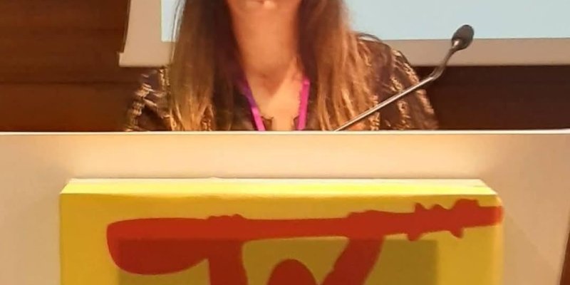 Kürsü arkasında konuşma yapan Rukiye Kara’nın portresi. Açık tenli, uzun düz saçlı (üst kısımlar koyu kahverengi, uçlara doğru daha açık/kumral tonlarda) ve gözlük takıyor. Gülümsüyor ve yüzü kameraya dönük. Önünde açık renkli bir kürsü; sağ tarafta ince saplı bir mikrofon. Kürsünün ön yüzünde sarı zeminli bir afişte kırmızı bir silüet, başının üstünde uzun bir nesneyi (kürek ya da sopa gibi) iki eliyle yukarıda tutuyormuş gibi görünüyor. Arka planda büyük, açık renkli bir projeksiyon/perde alan