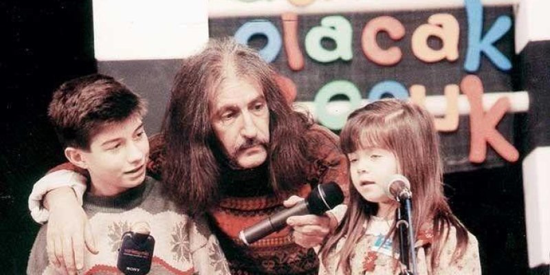 Bir sahnede iki çocukla birlikte duran uzun saçlı, bıyıklı Barış Manço. Koyu kahverengi uzun saçları omuzlarının altına kadar iniyor ve kalın bir bıyığı var; iki kolunu da çocukların omzuna dolamış. Sol tarafta kısa koyu saçlı bir erkek çocuk renkli desenli bir kazak giymiş ve elinde üzerinde “SONY” yazan bir mikrofon tutuyor; başını sağa çevirip bakıyor. Sağ tarafta uzun kahverengi saçlı, kaküllü bir kız çocuğu açık renkli, desenli bir üst giymiş; önünde ayaklı bir mikrofon var ve Manço elindek