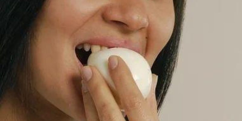Genç bir kadın, soyulmuş haşlanmış bir yumurtayı ağzına götürürken omuz hizasına kadar kadrajlanmış, başı hafif sağa dönük. Orta tenli, düz koyu renk saçlı ve kahverengi gözlü. Gözleri sağ tarafa doğru yönelmiş, yüzünde hafif bir gülümseme ve rahat bir ifade var. Sağ elinin parmaklarıyla beyaz yumurtayı tutuyor; yumurta dudaklarının arasında, ısırmak üzere. Dişleri hafifçe görünüyor. Üzerinde sade, kısa kollu beyaz bir tişört. Arka plan tek renk, açık bej tonunda.