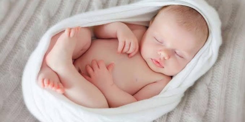 Beyaz bir örtüye sarılmış ve uyuyan bir bebek. Rahat bir şekilde uyurken elleri ve ayakları gevşek bir pozisyonda duruyor. Huzurlu ve sakin bir ifadeye sahip.