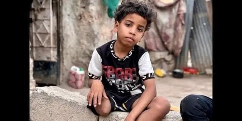 Beton bir basamak üzerinde oturan küçük bir çocuk. Kısa, dağınık siyah saçlı; koyu renk gözlü; orta-koyu tenli. Başını hafif yana eğmiş ve kameraya doğru ciddi ya da düşünceli bir ifadeyle bakıyor. Siyah ağırlıklı, kısa kollu bir tişört giymiş; üzerinde büyük kırmızı ve beyaz yazılar Siyah şort giymiş ve bacakları önde, dizleri bükülü şekilde oturuyor. Arka planda eski ve yıpranmış bir duvar, asılı duran kumaş parçaları, bir metal kapı/ızgara ve yerde bazı kaplar ile eşyalar. Sağ altta başka bir