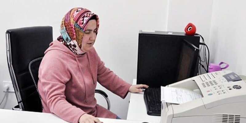 Bir ofis ortamında masada oturan başörtülü bir kadın pembe bir kapüşonlu üst giymiş, çok renkli desenli bir başörtüsü. Beyaz bir masanın arkasında oturuyor; sağ eliyle bilgisayar klavyesine, sol eliyle masadaki akıllı telefona uzanmış. Önünde büyük, açık renkli bir faks veya çok fonksiyonlu yazıcı ve üzerinde kâğıtlar, bilgisayar faresi ve küçük bir aygıt. Arka tarafta siyah kasalı bir masaüstü bilgisayar; kasanın üstünde kırmızı renkli, pelüş benzeri küçük bir nesne. Ortam aydınlık, duvar ve ma