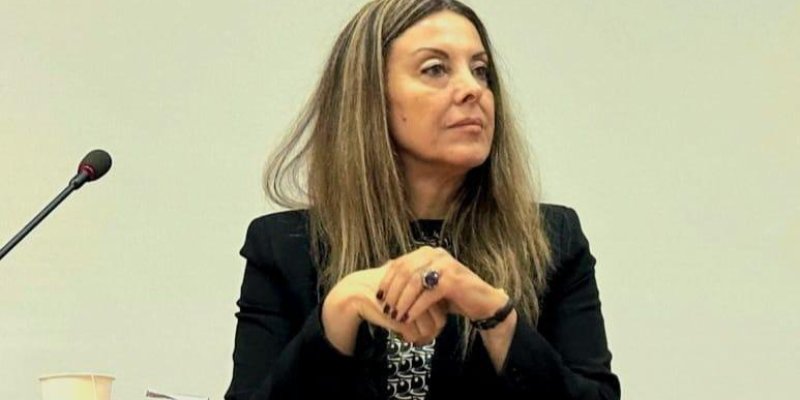 Orta yaşlarda, uzun açık kahverengi saçlı ve ciddi bir ifadeye sahip Neval Oğan Balkız, siyah bir ceket, desenli bir bluz giymiş ve bir toplantı veya konferans sırasında ahşap bir masanın arkasında oturuyor. Sağ eliyle sol bileğini tutuyor ve dikkatle bir noktaya bakıyor. Masanın üzerinde bir mikrofon ve bir kâğıt bardak bulunuyor.