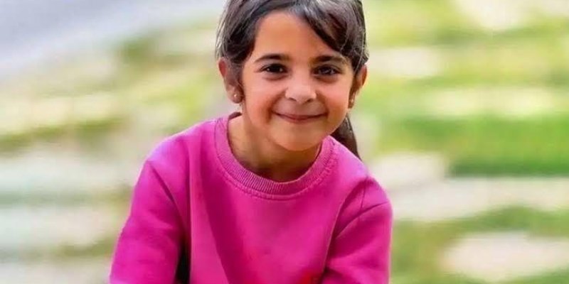 Pembe bir kazak giyen Narin gülümsüyor. Saçları koyu renkli ve arkadan toplanmış. Arka planda bulanık bir şekilde yeşil çimenler ve taşlar görünüyor. Yüz ifadesi neşeli ve mutlu.