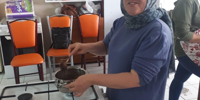 Turuncu sandalyelerin ve beyaz dolapların olduğu mutfakta mavi kazaklı genç bir kadın ocağın başında tencerede keyifle yemek pişiriyor.