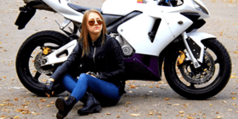Arkasında beyaz motosiklet olan bir kadın yerlerde sarı yapraklar olan bir asfaltın üzerinde oturmuş. Güneş gözlüklü ve kahverengi, düz uzun saçlı bir kadın, lacivert bir kot pantolon, siyah bot, siyah deri ceket giymiş. Ellerini dizlerinin üzerine koymuş.