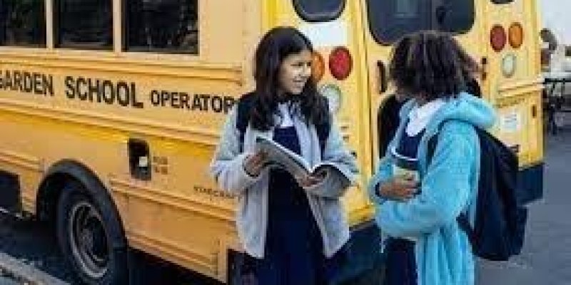Sarı öğrenci servisinin önünde, ellerinde kitap defter olan ve öğrenci forması giymiş iki kız öğrenci karşılıklı sohbet ediyor.