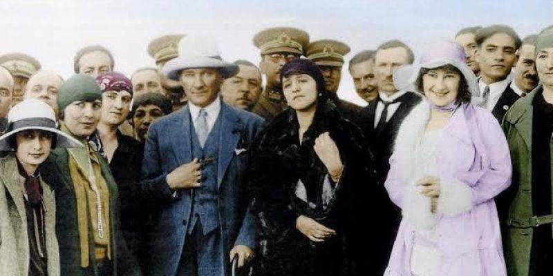 Mustafa Kemal Atatürk'ün bir grup insanla birlikte çekilmiş görüntüsü. Atatürk, fotoğrafın ortasında, açık renk bir takım elbise ve beyaz bir şapka giymiş olarak duruyor. Etrafında kadınlar ve erkekler bulunuyor. Kadınlar, dönemin modasına uygun şapkalar ve elbiseler giymişler. Erkeklerin bir kısmı askeri üniforma içinde, diğerleri takım elbise giymiş. Fotoğrafın arka planında daha fazla insan var ve hepsi Atatürk'ün etrafında toplanmış.