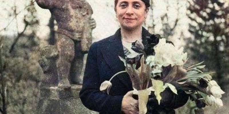 Safiye Ali açık havada bir heykelin yanında duruyor. Koyu renk bir ceket ve başına uygun bir şapka giymiş. Elinde büyük bir çiçek buketi tutuyor. Heykel, bir çocuğu tasvir ediyor ve bir kaide üzerinde duruyor. Arka planda ağaçlar ve doğal bir ortam görülüyor. Safiye Ali hafifçe gülümsüyor ve doğrudan kameraya bakıyor.