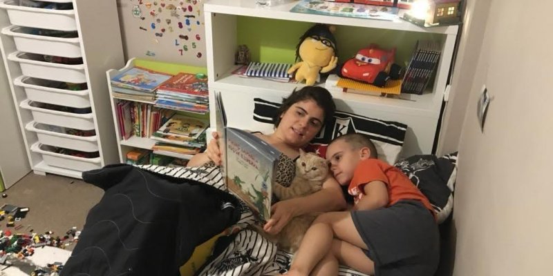 Yatakta yan yana uzanmış iki çocuk ve aralarında bir kedi. Daha büyük olan çocuk sırtüstü yatıyor, kucağında krem rengi, kısa tüylü bir kedi ve elinde resimli bir kitap tutuyor; sayfalar açık ve ikisinin de bakışı kitaba dönük. Küçük olan çocuk turuncu tişört ve gri şort giymiş, kıvrılmış şekilde ablasına/abisine sokulmuş, başı kedinin ve diğer çocuğun göğsüne yaslanmış. Yatak siyah beyaz desenli nevresimlerle kaplı. Başucunda beyaz bir raflı başlık; orta rafta yeşil arka fonda sarı saçlı, gözlü