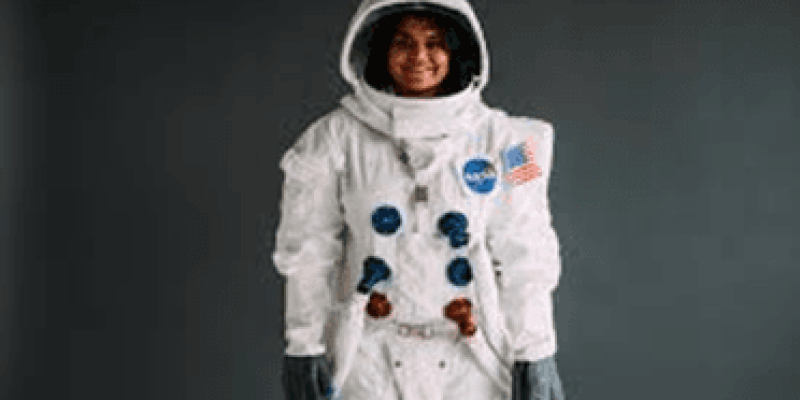 Gri bir arka plan önünde duran bir kadın astronotu gösteren bir fotoğraf. Astronot beyaz bir uzay giysisi giymiş ve başında beyaz bir kask var. Kaskın vizörü açık. NASA amblemi ve Amerikan bayrağı giysinin üzerinde yer alıyor. Astronotun yüzü gülümseyerek görünüyor ve eldivenli elleri yanlarda duruyor.