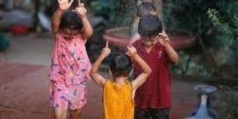 Yeşillikli, doğal ve yağmurlu bir ortamda pembe, sarı ve kırmızı kıyafetli üç çocuk ellerini havaya kaldırmış.