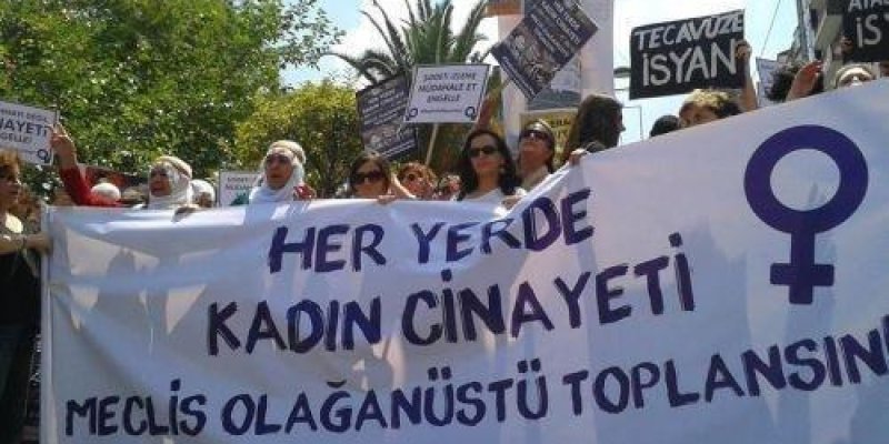 Kalabalık bir protesto gösterisinde önde taşınan büyük beyaz bir pankart. Pankartın sağ tarafında mor renkte kadın sembolü. Pankart üzerinde şu yazılar: “HER YERDE KADIN CİNAYETİ” “MECLİS OLAĞANÜSTÜ TOPLANSIN” “#KadınKatliamıVar”. Pankartın arkasında çoğu kadın olan bir grup, ellerinde dövizlerle duruyor. Dövizlerden birinde “TECAVÜZE İSYAN” yazısı seçiliyor. Açık havada, ağaçlar ve bir direk görünür durumda.