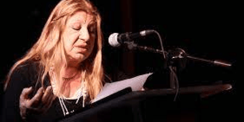 Ayla Algan, mikrofonlu bir kürsüde duruyor ve elinde kağıtlar tutuyor. Orta yaşlı ve sarı uzun saçlı. Ciddi bir ifadeyle kağıtlarına bakıyor ve konuşuyor.
