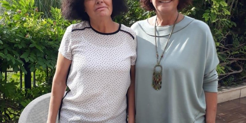 Açık havada, yeşilliklerle çevrili bir bahçede duran iki kadın. Soldaki kadın kısa kollu, beyaz ve siyah puantiyeli bir tişört giymiş, siyah saçları omuzlarının biraz altında. Sağdaki kadın, uzun kollu, açık mavi bir bluz giymiş, kısa kıvırcık kahverengi saçlı ve gülümsüyor. Önlerindeki kahverengi ahşap masada, büyük bir bez çanta ve bir tabakta hurmalar var. Arka planda çeşitli ağaçlar ve bitkiler görülüyor.