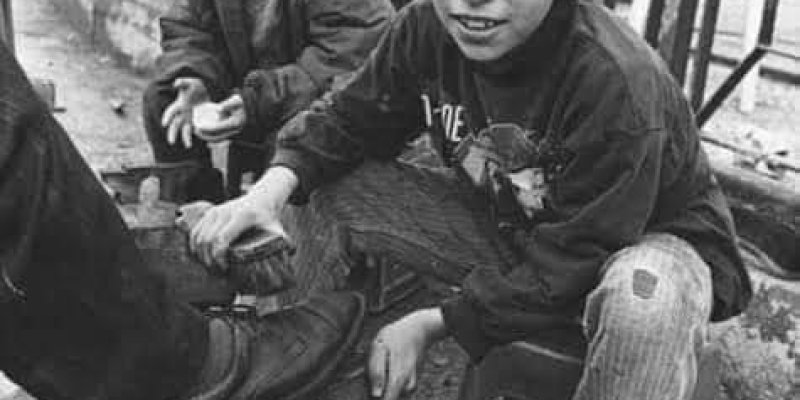 Sokakta ayakkabı boyacılığı yapan iki genç çocuk. Önde oturan, bir taburede ve bir müşterinin ayakkabısını boyuyor. Üzerinde koyu renkli bir kazak ve yırtık bir kot pantolon var. Yanında, başka bir taburede oturan ikinci bir çocuk gülümsüyor ve üzerinde mont benzeri bir giysi var. Arka planda, demir parmaklıklar ve ağaçlar görülüyor. Fotoğraf siyah-beyaz çekilmiş.
