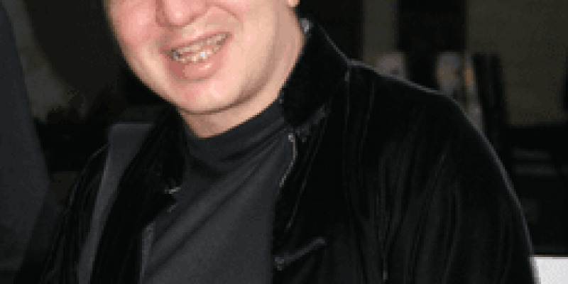Fazıl Say, siyah yakası dik kadifemsi bir ceket ve ceketinin içinde füme rengi hakim yaka hafif parlak bir bluz giymiş gülümsüyor.