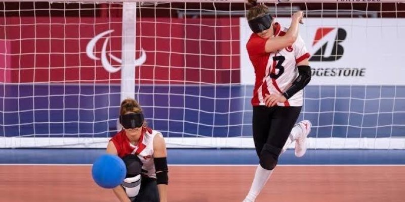Tokyo 2020 Paralimpik Oyunları'nda goalball oynayan iki kadın sporcu. Her iki sporcu da göz bandı takıyor. Sağdaki sporcu, 13 numaralı formayı giymiş ve topu atmak üzere. Sol taraftaki sporcu dizlerinin üzerinde oturuyor ve topu kontrol etmeye çalışıyor. Arka planda, oyun alanını çevreleyen bir ağ ve "TOKYO 2020" yazısı dikkat çekiyor. Zemin kırmızı renkte ve top mavi. Sporcuların formaları kırmızı ve beyaz renklerde.