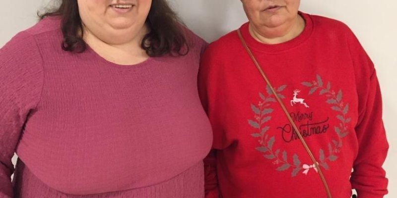 Kırmızı ve pembe bluzlu, spor giyimli genç ve orta yaşlı iki kadın gülümseyerek ayakta duruyorlar.