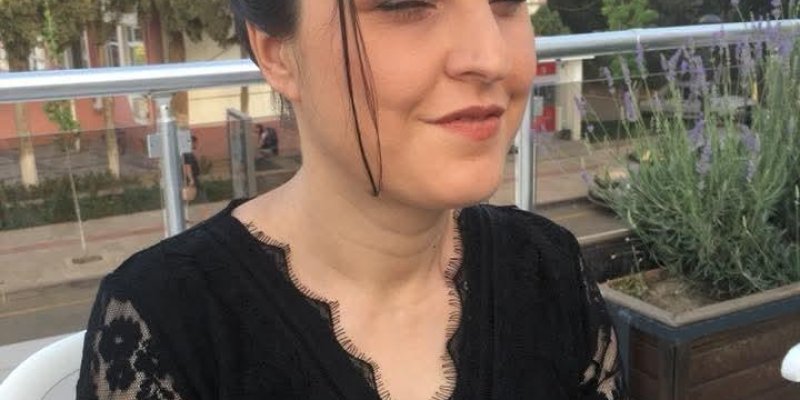 Siyah dantel detaylı elbise giymiş Hasibe Boztepe’nin saçları yukarıdan toplanmış ve önde birkaç tutam saç yüzüne düşüyor. Gözleri kapalı ve hafifçe gülümsüyor. Arka planda açık havada, çiçeklerin ve ağaçların olduğu bir ortamda oturuyor. Yanında beyaz plastik bir sandalye ve büyük bir saksıda lavanta benzeri mor çiçekler var. Gün ışığı ortamı aydınlatıyor.