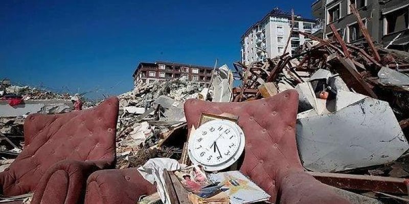 Yıkıntıların önünde iki bordo renkli, kapitone desenli koltuk. Koltukların arasında, üzerinde karışık kâğıtlar ve fotoğraflar bulunan ahşap görünümlü bir parça ve onun üzerinde beyaz kadranlı yuvarlak bir duvar saati duruyor. Arka planda büyük miktarda moloz, kırılmış beton parçaları ve bükülmüş metal iskeletler, sağ tarafta ağır hasarlı bir bina, daha uzakta ayakta duran birkaç apartman görülüyor. Gökyüzü açık ve mavi.