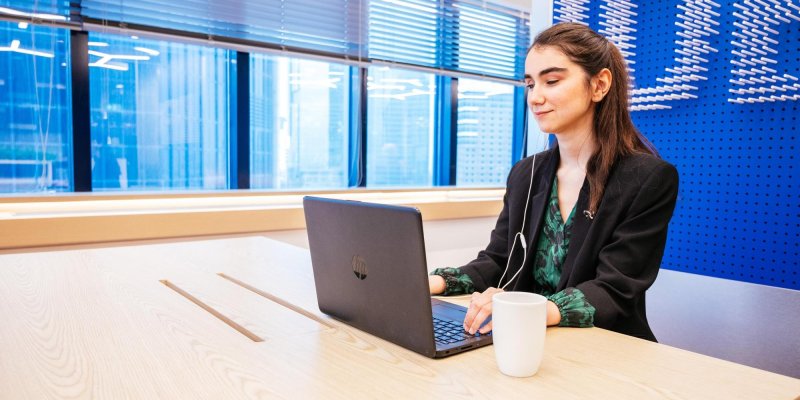 Genç bir kadın çalışma masasında laptop ile çalışıyor. Uzun kestane rengi saçlarını yarım toplamış, siyah ceket içine yeşil üzerine siyah desenli bluz giymiş. Tek kulağında kulaklık ve masada kupası var. Ofiste arka planda mavi zemin üzerine beyaz harfler var.