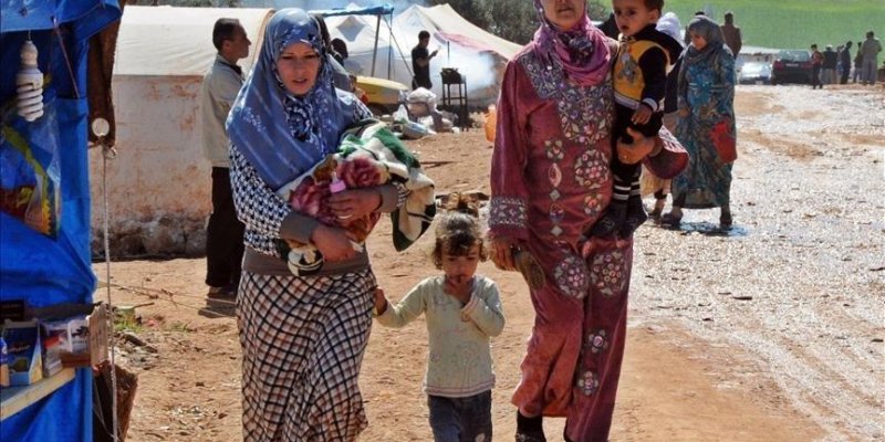 Açık havada, toprak zeminde kurulmuş çadırların arasında yürüyen insanlar. Ön planda iki yetişkin kadın ve iki küçük çocuk. Soldaki kadın, başı açık mavi tonlarında bir eşarp ve üstünde mavi-siyah desenli uzun bir elbise ile etek giymiş. Kucağında renkli battaniyelere sarılı bir bebek taşıyor. Yanında, elinden tuttuğu küçük bir kız çocuğu var; çocuk açık renk uzun kollu bir üst, koyu renk pantolon ve açık renk ayakkabılar giymiş. Sağdaki kadın, pembe tonlarında, üzerinde parlak işlemeler ve dese