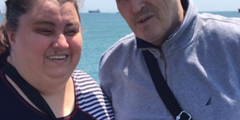 Deniz kenarında duran Lütfü Tokat ve eşi Selvet Bayraktar Tokat. Selvet solda, koyu renkli ve çizgili bir bluz giymiş, saçları toplu ve gülümsüyor. Lütfü sağda gri bir fermuarlı üst giymiş, omzunda çapraz bir çanta askısı bulunuyor ve hafif bir gülümsemeyle kameraya bakıyor. Arka planda mavi bir deniz ve ufukta birkaç gemi görülüyor.