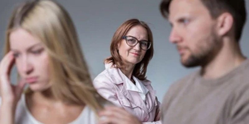 Üç kişiden oluşan bir grup, bulanık bir ön plan ve daha net bir orta planla gösteriliyor. Ön planda solda sarı saçlı bir kadın; başını hafif eğmiş, yüzünde üzgün ya da endişeli bir ifade ve eli şakağına yakın. Sağda, sakallı bir adam profilden görünüyor; yüzü net değil ama ifadesi ciddi ya da dalgın duruyor. Orta kısımda, bu iki kişinin arasında, gözlüklü orta yaşlı bir kadın net şekilde odakta; açık renk bir ceket giymiş, hafifçe gülümsüyor ve doğrudan karşıya bakıyor.