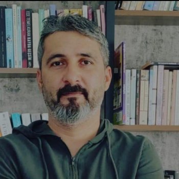 Gri saçlı ve sakallı, kollarını çaprazlayarak kameraya bakıyor. Üzerinde koyu yeşil bir kapüşonlu sweatshirt var. Arka planda, kitaplarla dolu bir kitaplık görülüyor. Kitapların bazıları dikey, bazıları yatay olarak yerleştirilmiş. Kitapların kapakları çeşitli renklerde ve arka plan duvarı gri tonlarında.