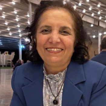 Mavi blazer ceket giymiş, koyu renk omuz hizasında dalgalı saçlı, kameraya dönük ve gülümsüyor. Ten rengi açık-orta tonda. İçeride, tavan boyunca küçük ışıklar bulunan bir mekânda oturuyor; arka planda masa-sandalye ve insanlar silik şekilde seçiliyor. Boynunda siyah ipli bir kolye ve yuvarlak koyu renk bir kolye ucu. Ceketin altında açık renk, desenli ve yakası fırfırlı bir bluz. Sol alt köşede “AI tarafından oluşturulan içerik” yazısı.