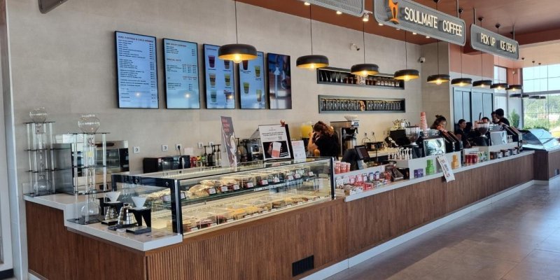 Geniş, modern tasarımlı bir kahve dükkânı tezgâhı. Önde uzun, L şeklinde beyaz tezgâhın ön yüzü ahşap kaplamalı. Tezgâhın sol kısmında camlı bir vitrin içinde sandviçler, kruvasanlar ve tatlılar sıralı; üst raflarda paketli ürünler, alt raflarda küçük kek ve pastalar. Vitrinin hemen yanında pour-over kahve demleme ekipmanları ve metal sürahiler duruyor. Tezgâhın orta bölümünde büyük bir meyve suyu veya limonata dispenseri, arkasında kahve makineleri ve çeşitli şurupların şişeleri. Hazırlık alanı