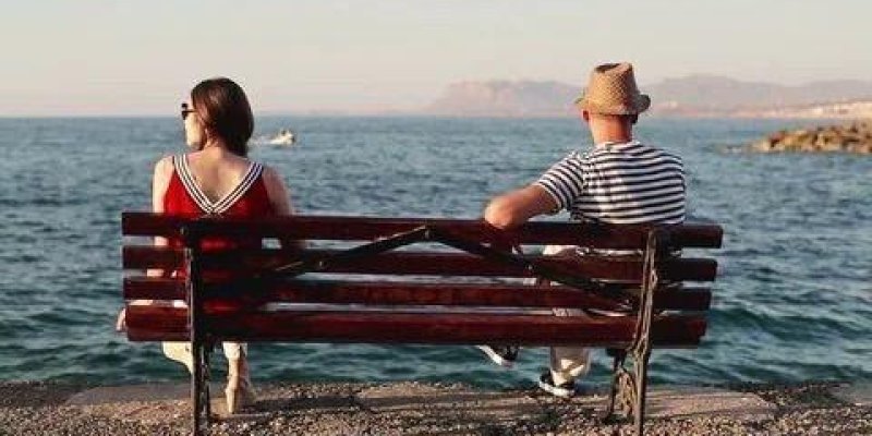 Deniz kıyısında ahşap bir bankta oturan iki kişi. Bank kadraja arkadan görünecek şekilde yerleştirilmiş; önlerinde sakin bir deniz ve uzakta dağ silueti. Solda omuz hizasında kısa koyu kahverengi saçlı, kırmızı elbiseli bir kadın oturuyor; elbisenin kol ve yaka kısmında beyaz şerit detayları. Sağda, başında hasır şapka olan bir erkek oturuyor; üstünde yatay lacivert-beyaz çizgili bir tişört ve açık renk pantolon var. Kadın sola doğru, erkek sağa doğru dönük; aralarında bir mesafe bulunuyor ve bi