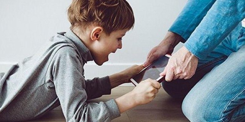 Yerde yüzüstü uzanan küçük bir çocuk ve dizlerinin üzerinde duran bir yetişkin. Çocuk gri, uzun kollu bir üst giymiş; kısa kahverengi saçlı ve ağzı açık şekilde heyecanlı gülüyor. Çocuğun iki eliyle tuttuğu dikdörtgen bir cihaz (tablet ya da büyük ekranlı telefon gibi) yetişkinin ellerine doğru uzanıyor. Yetişkin mavi kot pantolon ve mavi uzun kollu bir üst giymiş; iki eliyle cihazı tutuyor ya da cihazın kenarına dokunuyor. Zemin ahşap görünümlü; arka tarafta açık renk bir duvar var.