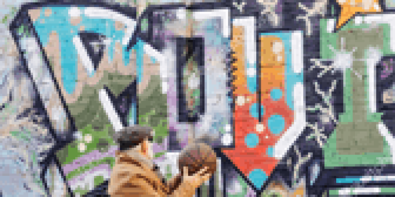 Büyük puntolarla yazılmış yazıların olduğu büyük bir duvarda kırmızı çizgilerle çizilmiş bir basketbol potası, potanın önünde tekerlekli sandalyede yaşlı bir adam var. Adam turuncu renkte pardösü, siyah pantolon ve kahverengi ayakkabı giymiş, kahverengi bir şapka takmış. İki eli havada turuncu basketbol topu tutuyor.