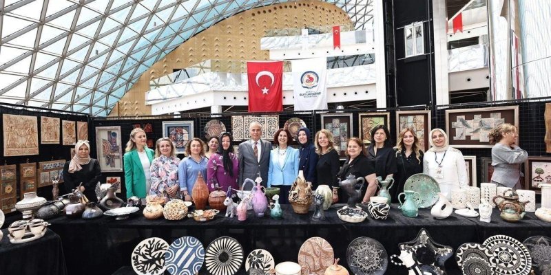 Bir sergi standının önünde yan yana dizilmiş bir grup yetişkin insan ve önlerinde sergilenen el yapımı seramik/çini eserler. Fotoğraf kapalı bir alanda çekilmiş; üstte camdan kubbe/çatı yapısı. Önde uzun bir masa boyunca çok sayıda seramik obje, tabaklar, vazolar, sürahiler, çaydanlık benzeri formlar, kaseler ve dekoratif figürler. Renkler ağırlıkla siyah, beyaz, turkuaz, yeşil, kahverengi ve mor; bazı tabaklarda siyah-beyaz geometrik desenler ve çizgisel motifler bulunuyor. Masanın ön kısmında