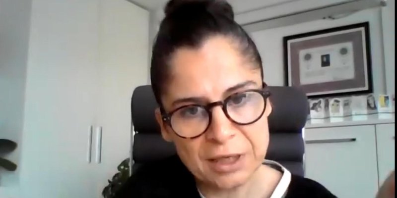 Konuğun fotoğrafı. Solda beyaz dolap, arkada diploma çerçevesi, siyah koltukta konuk oturuyor. Saçları kahverengi tepeden topuz yapılmış, gözünde siyah çerçeveli gözlük, ekrana bakıyor, konuşur vaziyette ağzı açık.