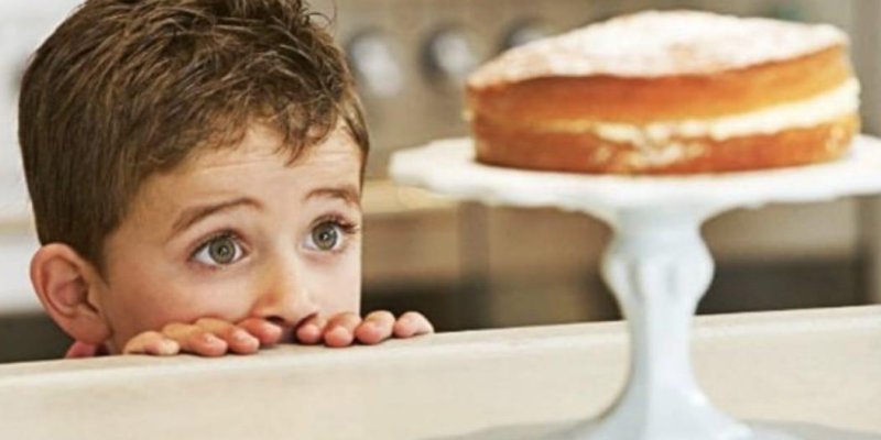 Bir tezgâhın arkasından gözleri görünen küçük bir çocuk. Tezgâhın üst kenarına iki eliyle tutunmuş, gözlerini sağ taraftaki pastaya doğru dikmiş. Sağda beyaz bir ayaklı servis tabağının üzerinde, ortasında beyaz kremalı katman olan yuvarlak bir kek duruyor; üstüne pudra şekeri serpilmiş gibi görünüyor. Arka plan bulanık bir mutfak görüntüsü.