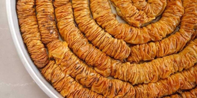 Yuvarlak bir tepsi dolusu ince kıvrımlı tatlı. Tepsinin içini tamamen kaplayan, ortadan başlayıp dışa doğru devam eden spiral şeklinde yerleştirilmiş uzun, rulo hamur şeritleri. Hamurlar çok katlı, sık sık pile yapılmış, büzgülü duruyor. Renkleri altın kahverengi ile koyu bal rengi arasında, üzerleri parlak. Spiral halkalar birbirine çok yakın, neredeyse aralarında boşluk yok. Tepsinin kenarı metalik, açık gri renkte ve sade. Aralarda hafifçe daha koyu kızarmış, yer yer küçük yanık noktalar bulu