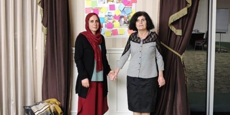 Söyleşi yapan iki kadın el ele tutuşmuş duruyor. Sol taraftaki kadın kırmızı bir eşarp, siyah üst ve kırmızı etek giymiş. Sağ taraftaki kadın siyah ayakkabılar, siyah etek ve siyah beyaz desenli bir gömlek giymiş. Arka planda bej renkli çizgili duvar kâğıdı ve iki adet koyu kahverengi perde görülüyor. Perdelerin arasında bir poster asılı ve üzerinde renkli not kâğıtları yapıştırılmış.