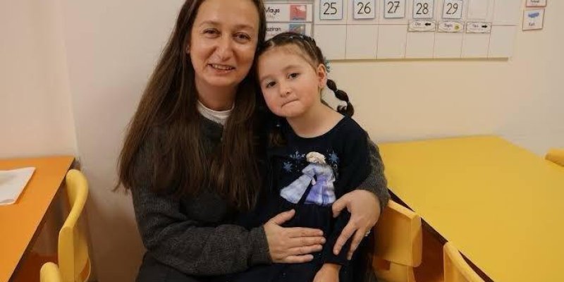 Bir kadın ve küçük bir kız çocuğu sarı masaların bulunduğu bir sınıfta oturuyor. Kadın, uzun kahverengi saçlı ve gri bir kazak giymiş. Kız çocuğu Elsa karakterinin resminin olduğu koyu renkli bir elbise giymiş ve saçları iki yandan örgülü. Arka planda, duvarda bir takvim var ve üzerinde günler ve aylar yazılı. Masalar ve sandalyeler sarı renkte. Kadın, kızı kucağına almış ve ikisi de kameraya bakarak gülümsüyor.