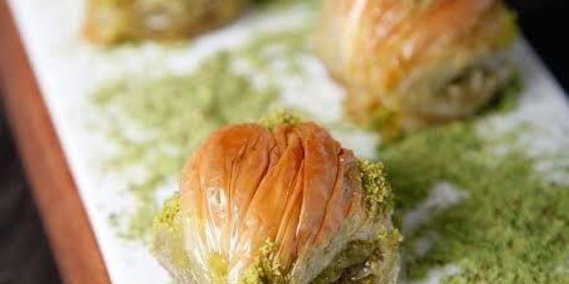 Beyaz bir tabak üzerinde dört adet baklava dilimi yer alıyor. Baklavalar, ince kat kat açılmış hamurdan yapılmış ve üzerleri altın rengi bir parlaklıkla kızarmış. Her bir baklava diliminin etrafında ve tabakta bol miktarda yeşil toz fıstık serpilmiş. Tabak, ahşap bir servis tahtası üzerinde sunulmuş. Arka planda bulanık bir şekilde başka baklava dilimleri ve fıstıklar görünüyor.