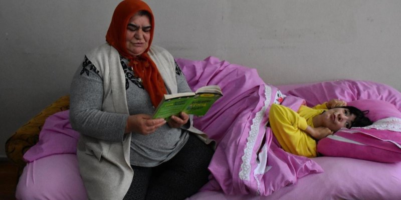 Pembe çarşaflı geniş bir yatakta yatan çocuk ve yanında oturan bir kadın, solda yatağın kenarında oturuyor. Kafasında turuncu eşarp, üzerinde gri uzun kollu bir bluz ve açık renk, cepli bir yelek; koyu renk desenli bir pantolon giymiş. Ellerinde açık yeşil kapaklı bir kitap tutuyor ve kitaba dönük, okur durumda. Çocuk sağ tarafta, pembe yorgan ve yastıkların arasında sırtüstü yatıyor. Üzerinde parlak sarı, uzun kollu bir üst. Başını hafif yan tarafa çevirmiş, bir eli başının altında, diğer eli ç