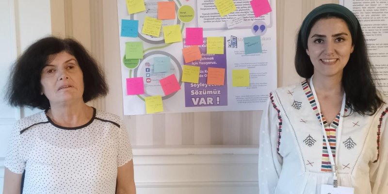 İki kadın renkli postitlerle donatılmış "Umudun Kadınları " afişinin iki yanında bulunuyor. İkisi de mavi kot pantolon üzerine beyaz bluz giymiş.
