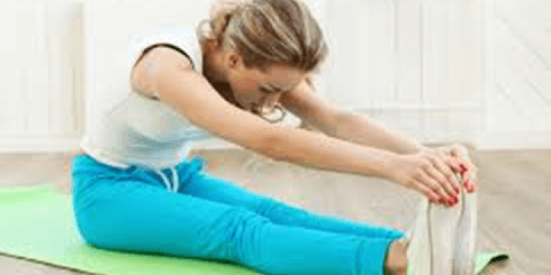 Bir kadın yeşil bir yoga matı üzerinde oturuyor ve esneme hareketi yapıyor. Bacaklarını düz bir şekilde öne uzatmış ve elleriyle ayak parmaklarına dokunmaya çalışıyor. Üzerinde beyaz bir tişört ve mavi bir eşofman altı var. Saçları arkadan toplanmış. Arka planda beyaz bir duvar ve ahşap zemin görülüyor.