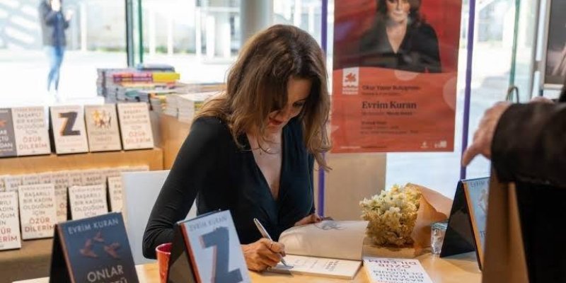 Kitap imza etkinliğinde bir kadın, masada oturmuş ve bir kitabı imzalıyor. Kadının önünde birkaç kitap yığılmış. Masanın üzerinde "Evrim Kuran" ismi yazılı kitaplar ve bir buket çiçek var. Arka planda, kadının büyük bir posteri asılı. Posterde "Evrim Kuran" ismi ve "Onlar Göçtü Buradan" kitabının tanıtımı yer alıyor. Çevrede başka kitaplar da sergilenmiş.