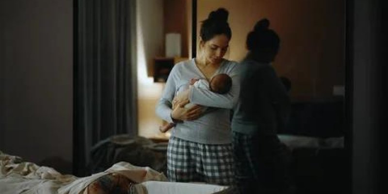 Loş ışıkta ayakta duran bir kadın, kucağında bir bebek tutuyor. Kadın gri uzun kollu bir üst ve kare desenli pijama altı giymiş. Saçları tepede dağınık bir topuz şeklinde toplanmış. Bebeği göğsüne yakın şekilde, koruyucu bir pozda kavramış; başı bebeğe doğru eğik, ifadesi yorgun ama şefkatli.