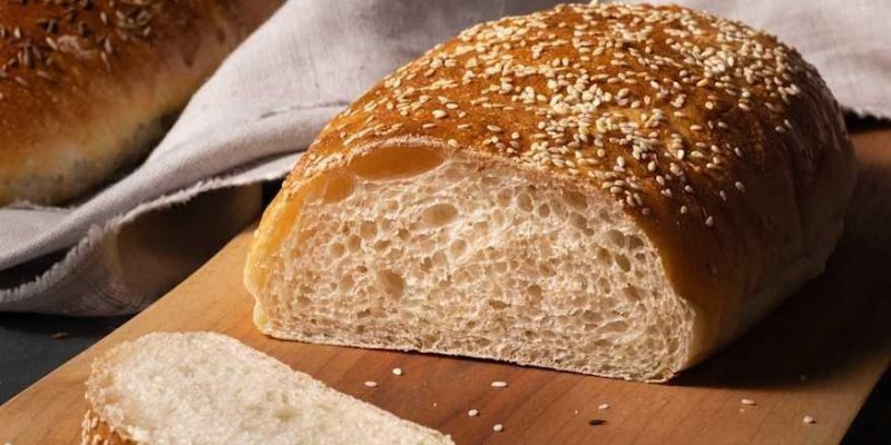 Susamlı ekmek somunu Tahta bir kesme tahtasının üzerinde, üstü susam taneleriyle kaplı altın-kahverengi kabuğa sahip parlak bir ekmek somunu duruyor ve somunun bir ucu ortadan kesilmiş, iç kısmı açıkta. İç dokusu açık renkli, yumuşak ve gözenekli. Somunun önünde, aynı ekmekten kesilmiş ince bir dilim tahtaya yatay şekilde yerleştirilmiş; kenarlarında ve üstünde birkaç susam tanesi. Arka planda açık renkli bir kumaş parçası ve bulanık görünen başka ekmek somunları.