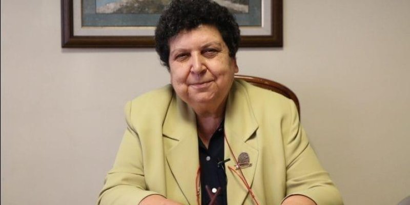 Kısa kıvırcık koyu saçlı Şenal Saruhan, bir masanın başında oturuyor. Yüzünde hafif bir gülümseme ve doğrudan kameraya bakıyor. Üzerinde açık sarı-krem tonlarında bir ceket, içinde koyu renk bir bluz. Ceketin ön kısmında bir rozet takılı ve boynundan aşağı doğru ince bir zincir veya kordonla bağlı bir gözlük sarkıyor; gözlük çerçevesi kırmızımsı renkte. Elleri masanın üzerinde.