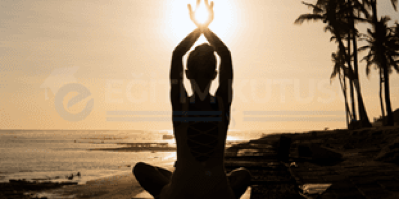 Güneşin batmak üzere olduğu bir sahil kenarında, palmiye ağaçlarının silüetleri arasında oturarak yoga yapan bir kişinin silüeti, ellerini başının üzerinde birleştirmiş. Deniz kenarında kayalık bir zemin üzerinde ve etrafında huzurlu bir atmosfer hâkim.